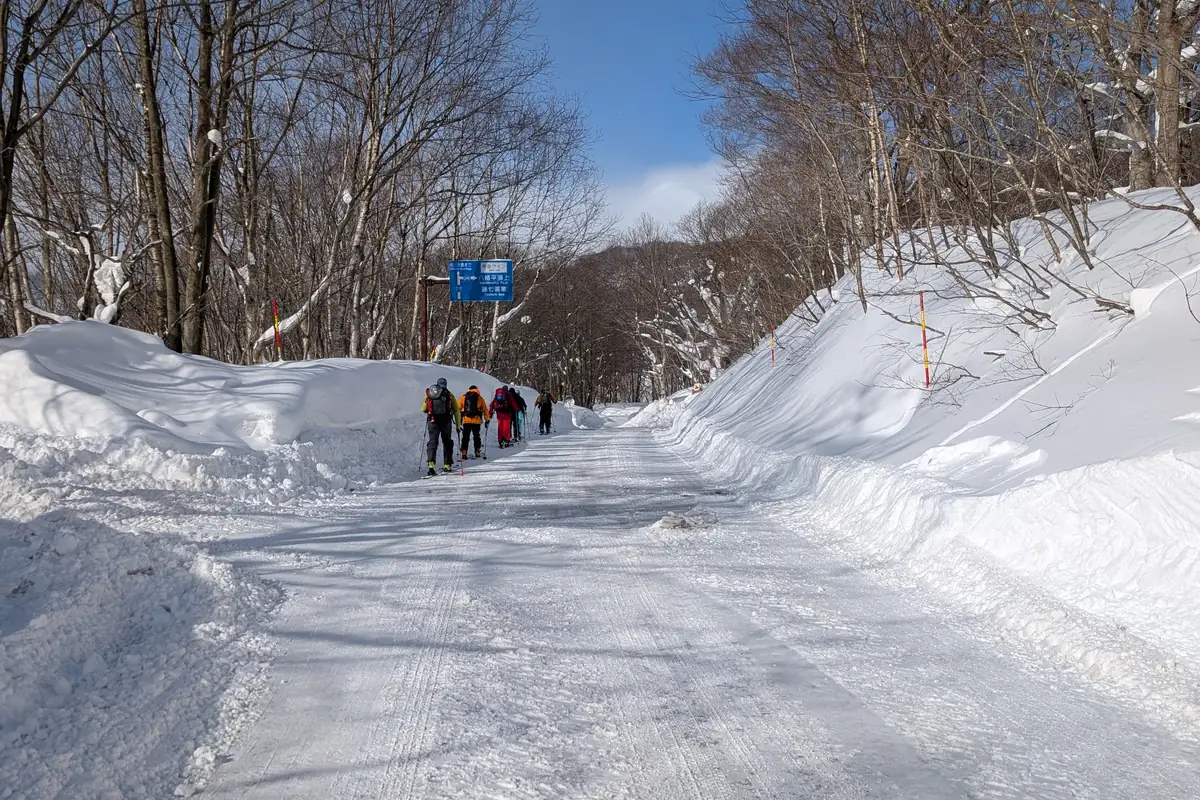 Ski tourers skinning up a snow-walled road toward the Hachimantai summit sign