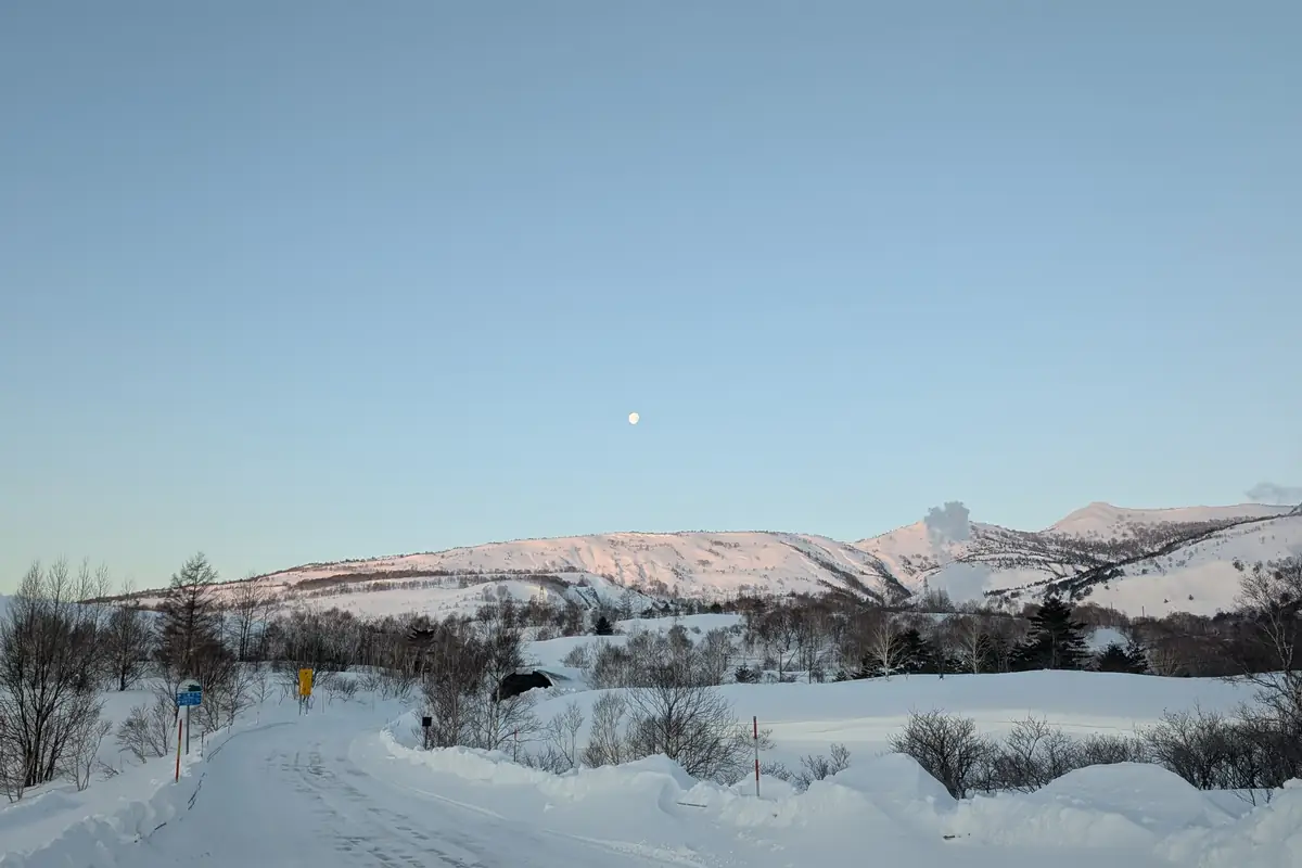 Snow-covered road at dawn with the moon above alpenglow ridges