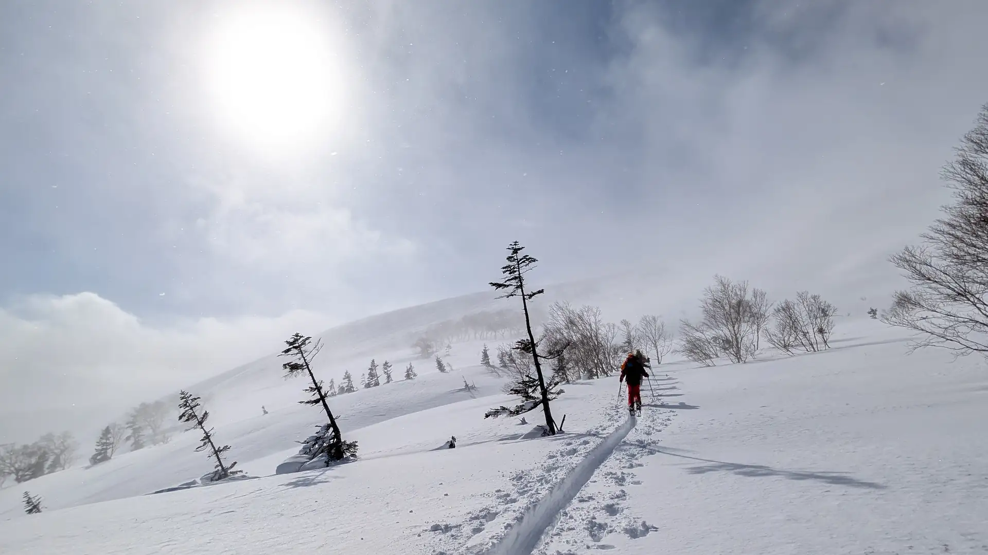 A skin track rising through mist toward a lone ski tourer below the sun